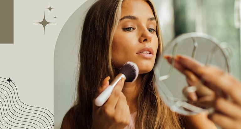 Young woman applying powder foundation with a makeup brush while looking into a handheld mirror.