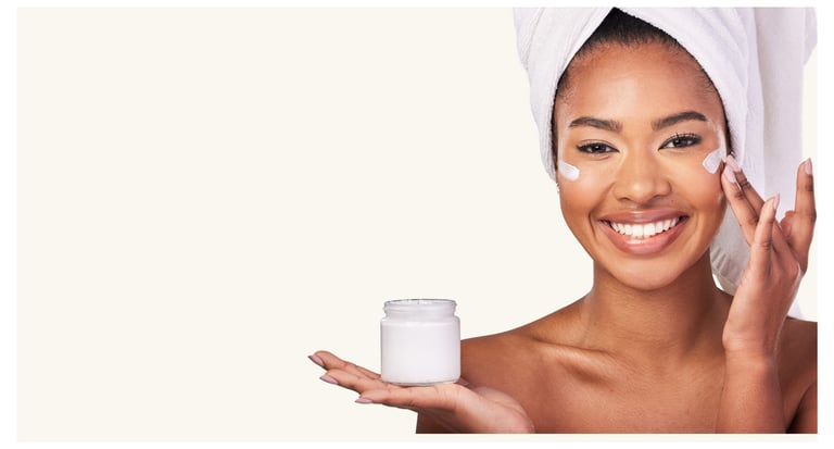 Smiling woman with a towel head wrap applying moisturizing face cream from a white skincare jar.