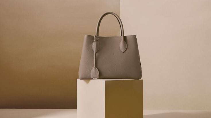 Elegante bolso de mano de piel color topo exhibido sobre un pedestal minimalista.