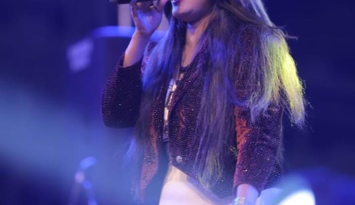 Female vocalist with long hair singing into a microphone during a live concert performance with stage lighting.