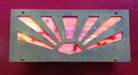 Brass sconce with colorful glass behind sunbust cutout.