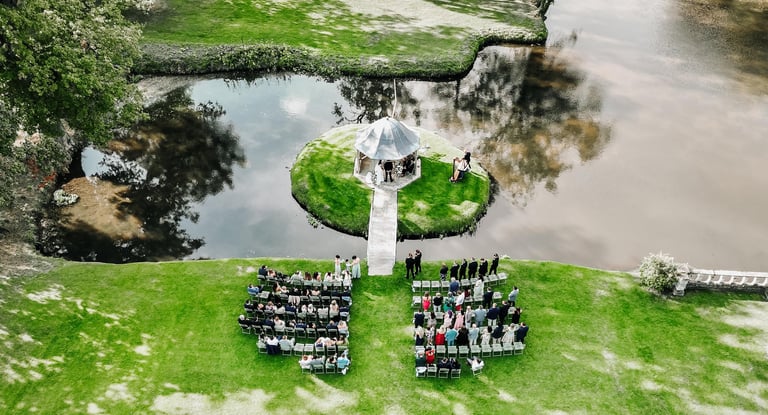Drone photo of an outdoor wedding ceremony by a lakeside gazebo surrounded by guests