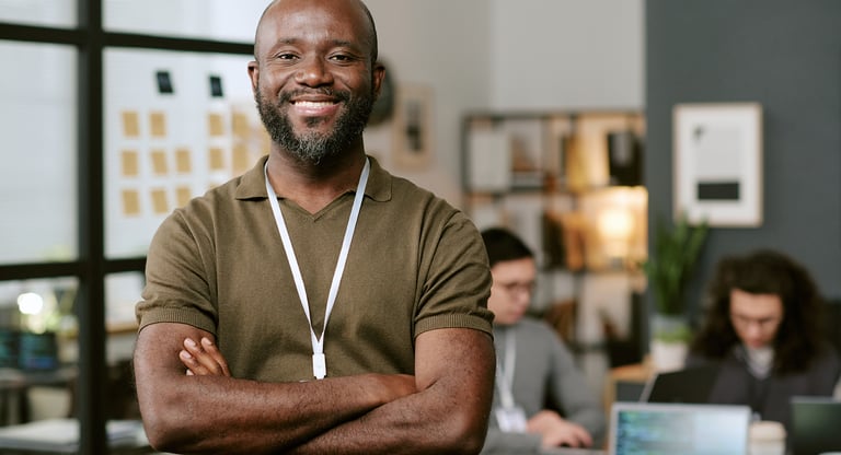 A smiling Black professional male standing with arms crossed in a modern open-plan office.