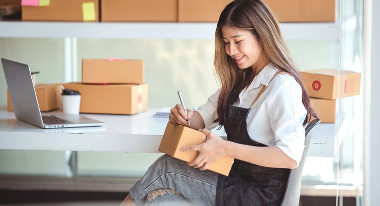 Smiling small business owner labeling cardboard shipping boxes for e-commerce order fulfillment.
