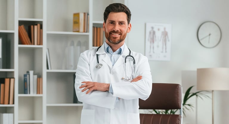 A smiling male doctor in a white lab coat with a stethoscope standing in a modern medical office.