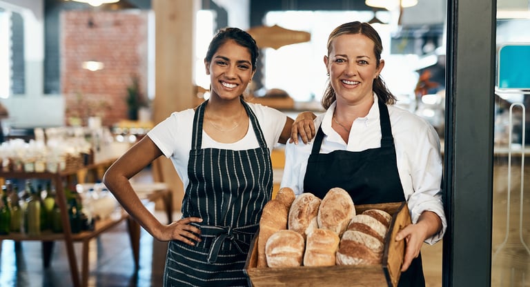 Two smiling female bakers holding a wooden crate of fresh artisanal bread loaves in a bakery.