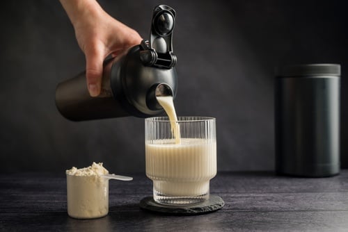 Hand pouring a vanilla protein shake from a black shaker bottle into a ribbed glass with supplement powder.