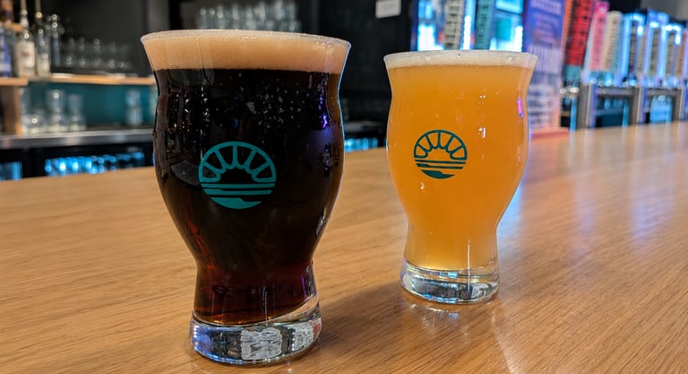 A dark stout and a hazy IPA craft beer served in tulip glasses on a wooden bar counter.