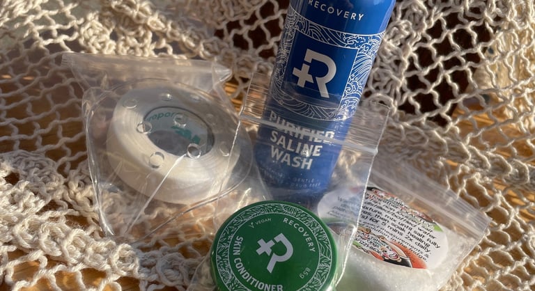 Piercing aftercare kit flatlay: Recovery brand saline spray & skin salve, protection patch + medical tape, & bag of sea salt.