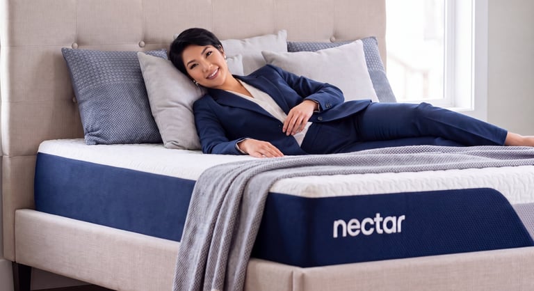 A smiling woman in a navy suit relaxes on a comfortable Nectar memory foam mattress with plush pillows.