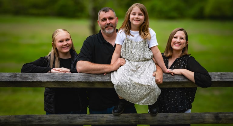 A happy family of four poses for a portrait outdoors behind a rustic wooden fence in a lush green field.