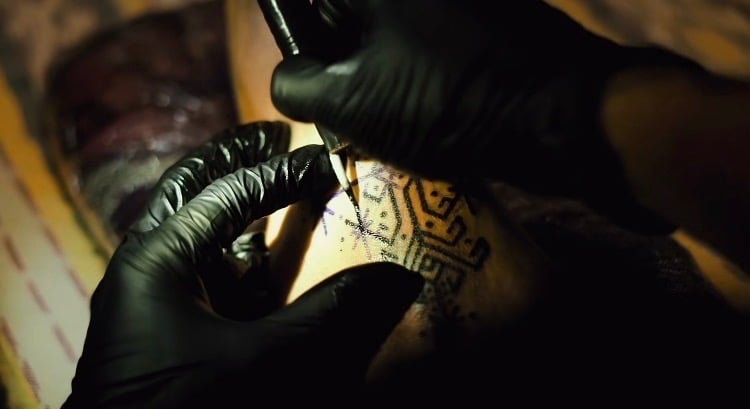 Professional tattoo artist in black gloves using handpoke technique to ink a geometric pattern.