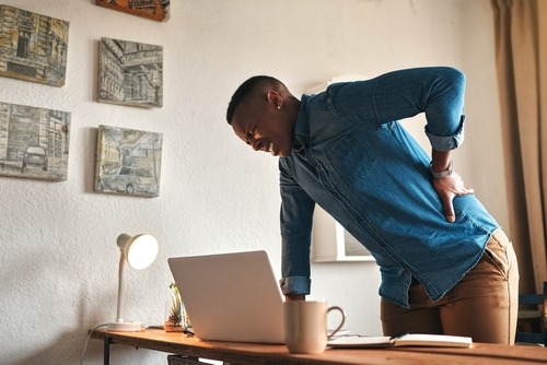 A man standing at a desk holding his lower back in pain while working on a laptop at home.