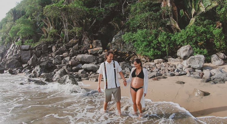 una pareja en la playa, la mujer está embarazada, se observa vegetación en la parte trasera