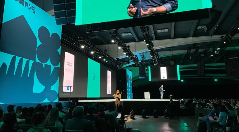 large conference audience facing an ASL interpreter standing under a large screen with the speaker