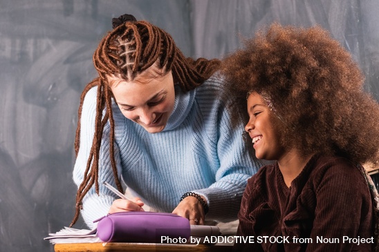Smiling woman helping a young girl with schoolwork in a warm, supportive setting.
