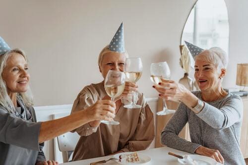 Tres mujeres mayores con sombreros de fiesta haciendo un brindis con vino en una celebración