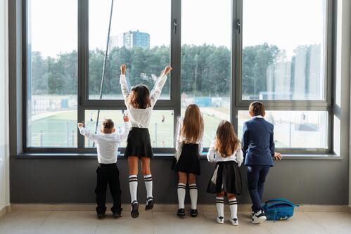 Estudiantes con uniformes escolares mirando por un gran ventanal hacia un patio de recreo