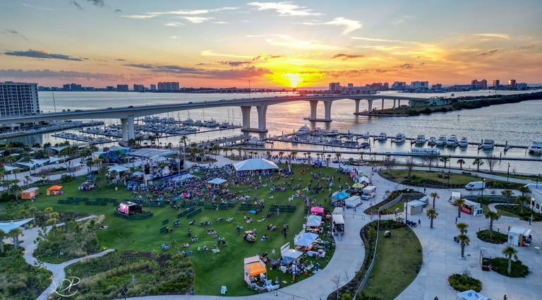 Aerial view of a waterfront park festival with a sunset backdrop over a bridge and marina.