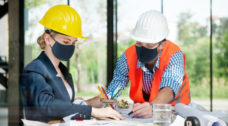 Two construction engineers wearing hard hats and face masks reviewing blueprints and architectural plans.