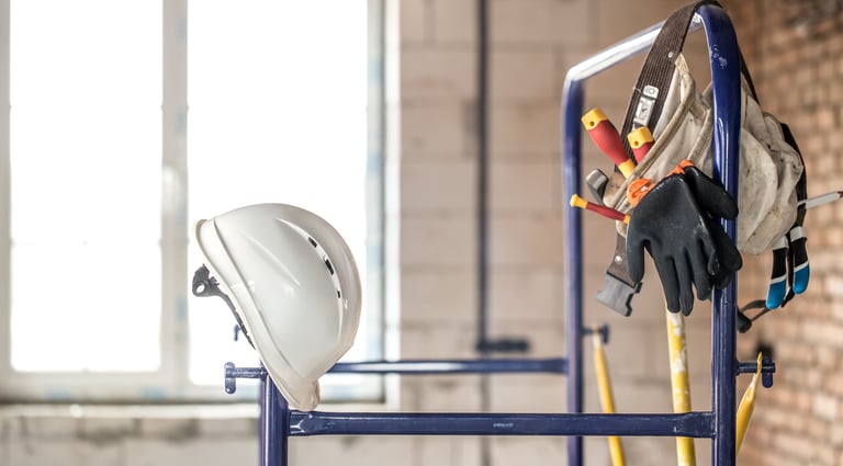 White construction hard hat and tool belt with gloves hanging on blue scaffolding at a job site.