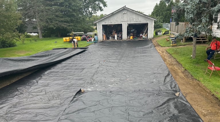 Workers laying black geotextile fabric for driveway stabilization in front of a residential garage.