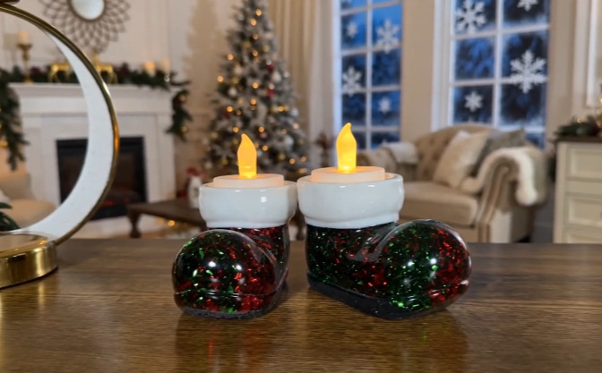 Two lighted Santa boot tea light candle holders on a table with a blurred Christmas tree background.