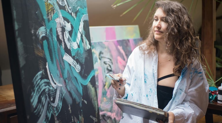 ovem mulher com camisa salpicada de tinta pintando em uma tela escura com pinceladas verde-azuladas.