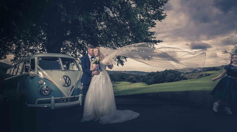 Bride and groom embrace by a vintage blue VW camper van with a flowing wedding veil in the wind.
