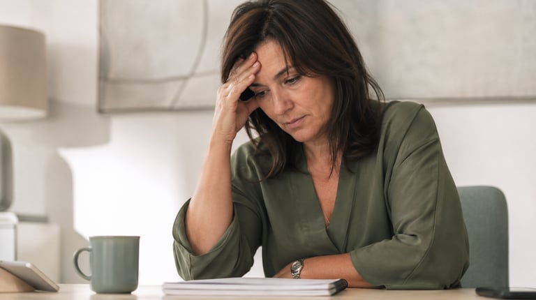 A stressed middle-aged woman sitting at a desk and holding her head while looking at documents.