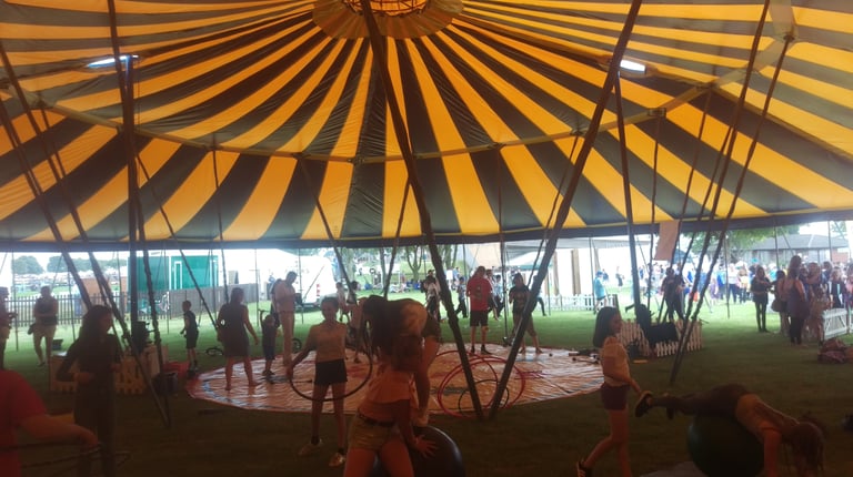 Children participating in a circus skills workshop inside a yellow and black striped marquee tent..