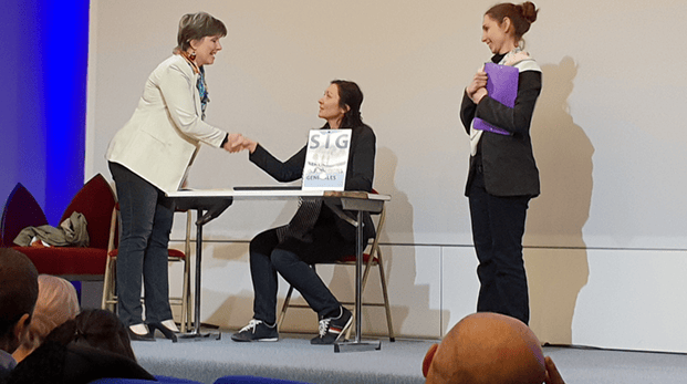 Sur scène une femme debout serre la main sous le regard d'une troisème forum accueil usager