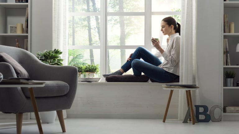 Habitación relajante, luminosa, pintada de blanco, con sillón gris y mujer sentada junto al ventanal