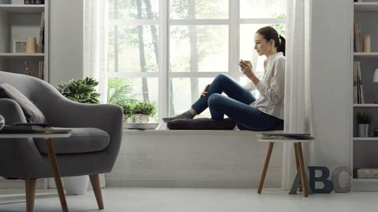 Habitación relajante, luminosa, pintada de blanco, con sillón gris y mujer sentada junto al ventanal