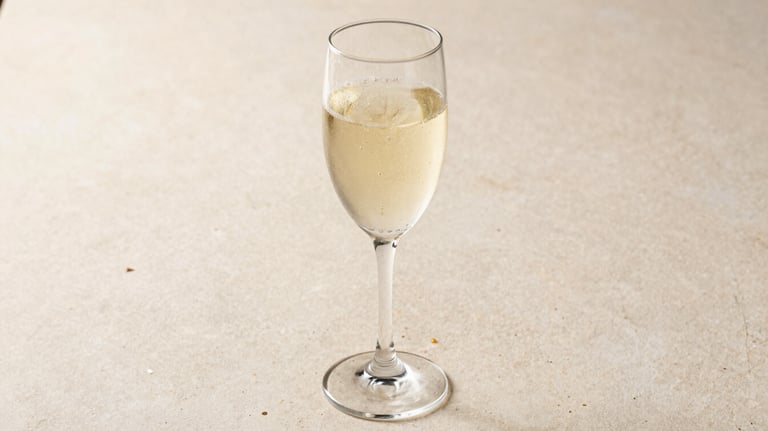 A crystal glass of sparkling wine with rising bubbles, set against a light beige stone surface. The lighting is bright and clean, typical of a refined Brazilian afternoon setting.