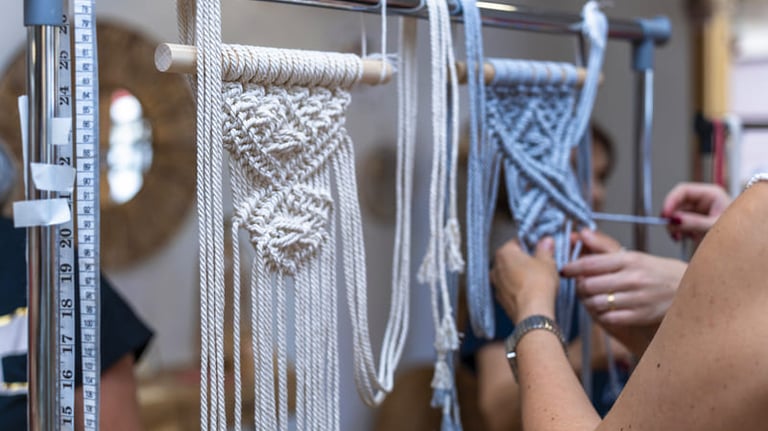 manos de 2 mujeres tejiendo macrame en bastidor de tono azul y tambien un tejido en tono blanco