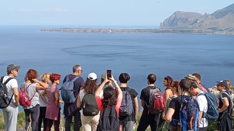 A group of hikers with backpacks overlooks the scenic blue coastline and mountains in Sicily.