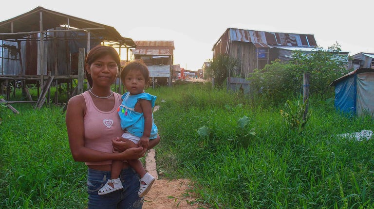 A smiling young woman holds a toddler in a rural village with wooden stilt houses and lush green grass.