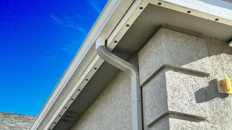 White aluminum seamless rain gutter and downspout installed on a stucco home exterior with a blue sky background.