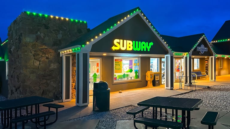 Subway sandwich shop exterior at night with green neon lights and outdoor patio seating.