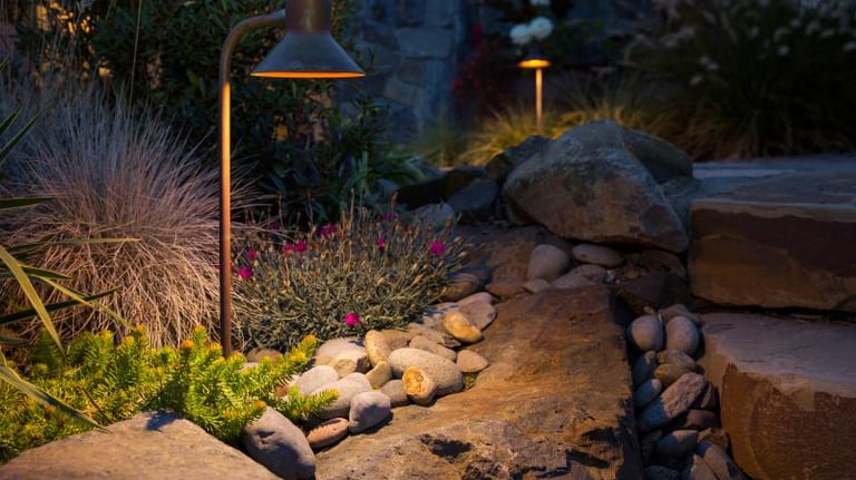 Warm outdoor lighting illuminates a stone pathway and garden plants, enhancing the nighttime landscape design.