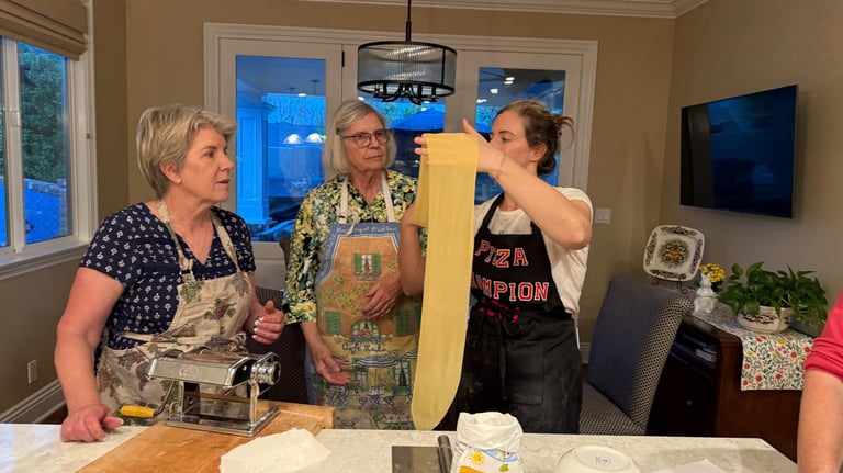 Three women wearing aprons make fresh pasta by hand using a stainless steel pasta roller machine in a home kitchen.
