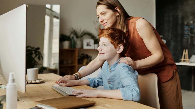 Lifestyle photo of woman standing behind son at desk in front of computer screen and keyboard.
