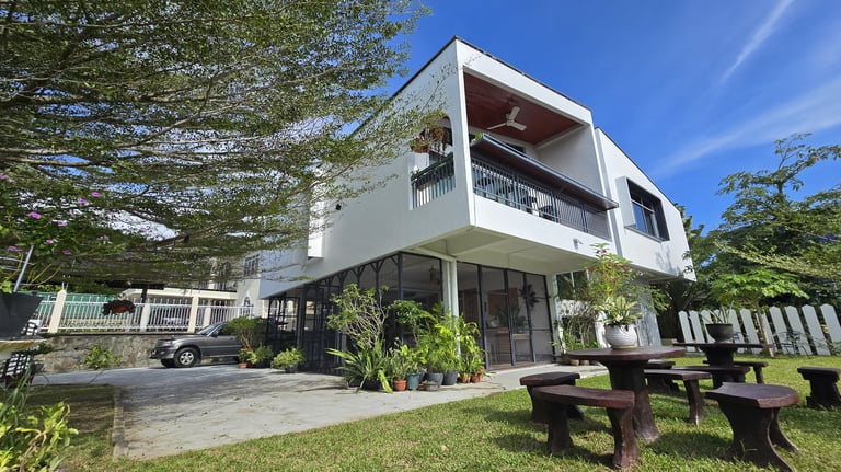 Modern white two-story house with a balcony, garden patio furniture, and lush green trees.