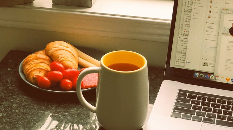 A remote work setup featuring a laptop, a mug of coffee, and a breakfast plate with croissants.