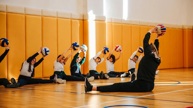A group of young volleyball players sit on the gym floor holding balls overhead while stretching, le