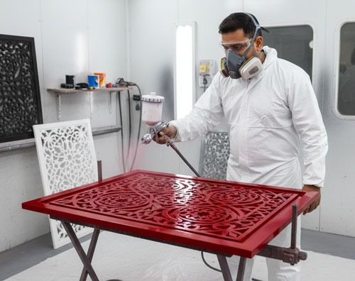 Professional painter in protective gear using a spray gun to apply red finish to an intricate laser-cut wood panel.