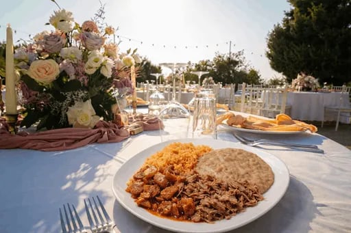 Plato con barbacoa, carnitas, arroz y frijoles sobre mesa elegante con centro de flores en boda al atardecer.