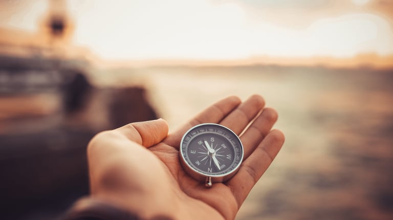 Hand holding a silver pocket compass against a blurred sunset horizon to find his way.