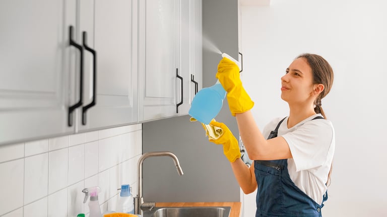 Pracownica firmy sprzątającej w rękawiczkach spryskuje środkiem czyszczącym szafki kuchenne nad zlewem